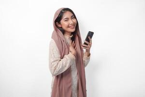 contento memore grato giovane asiatico musulmano donna mano su il petto sorridente mentre Tenere smartphone, isolato su bianca sfondo foto