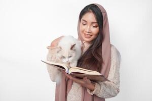 attraente giovane asiatico musulmano donna nel velo hijab sorridente mentre abbracciare un' bianca bambola di pezza gatto animale domestico e Tenere al Corano su il altro mano foto