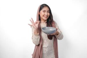 allegro asiatico musulmano donna nel foulard velo hijab Tenere ciotola con copia spazio dentro mentre fabbricazione un va bene cartello gesto. Ramadan concetto foto