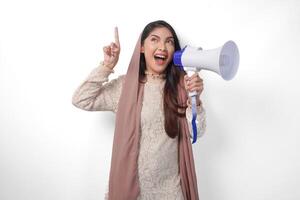 giovane asiatico musulmano donna indossare foulard velo hijab urlando a megafono mentre puntamento su per il copia spazio verso l'alto, isolato su bianca sfondo studio. Ramadan e eid mubarak concetto. foto