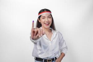 giovane asiatico donna con orgoglio mostrando poco dito immerso nel viola inchiostro dopo voto per Presidente e parlamento elezione, esprimendo eccitazione e felicità, indossare bandiera fascia per capelli e bianca camicia foto