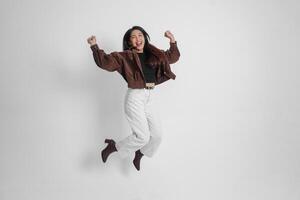 attivo asiatico ragazza è salto e raccolta mani su indossare Marrone giacca, isolato bianca colore sfondo foto