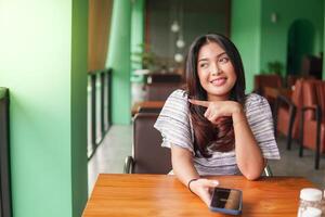 giovane bella asiatico donna seduta a un' ristorante per avere prima colazione mentre utilizzando smartphone con dito puntamento per sua giusto lato foto