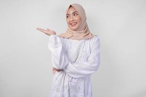 eccitato asiatico donna indossare hijab e bianca vestito puntamento per il copia spazio su sua giusto lato, in piedi al di sopra di isolato bianca sfondo. Ramadan e eid mubarak annuncio pubblicitario concetto foto