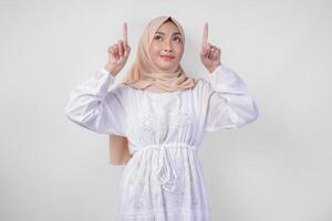 contento asiatico donna indossare hijab e bianca vestito puntamento su per il copia spazio sopra, in piedi al di sopra di isolato bianca sfondo. Ramadan e eid mubarak annuncio pubblicitario concetto foto