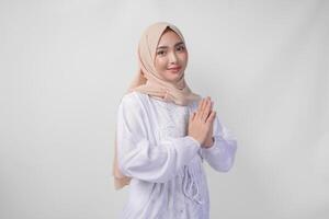 bellissimo asiatico musulmano donna indossare bianca vestito e hijab sorridente mentre fare formale benvenuto o saluto gesto, in piedi al di sopra di isolato bianca sfondo foto