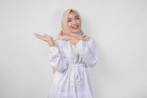 sorridente asiatico donna indossare hijab e bianca vestito puntamento su per il copia spazio su sua giusto lato, in piedi al di sopra di isolato bianca sfondo. Ramadan e eid mubarak annuncio pubblicitario concetto foto
