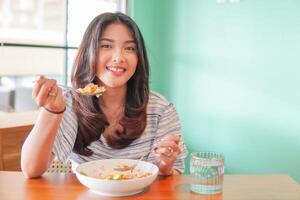 ritratto di un eccitato giovane asiatico donna indossare vestito seduta a un' ristorante, mangiare e godendo prima colazione con un' allegro Sorridi foto