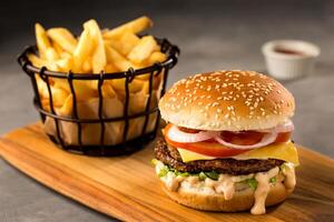 fresco speziato Manzo hamburger con francese patatine fritte benna, sesamo semi, pomodoro, cipolla, formaggio fetta, Maionese e ketchup servito su di legno tavola superiore Visualizza di Rifiuto veloce cibo foto