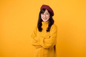 ritratto allegro giovane 30s asiatico donna indossare giallo maglione, rosso cappello e bicchieri mentre attraversamento braccio su il petto isolato su giallo sfondo. giovane talento artista stilista Guarda concetto. foto