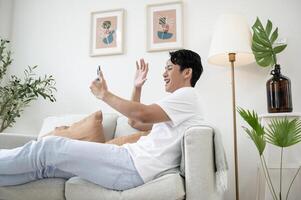 contento asiatico uomo seduta su divano utilizzando smartphone nel vivente camera a casa, rilassare tempo e stile di vita concetto foto