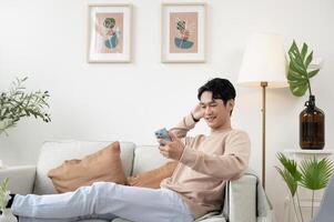 contento asiatico uomo seduta su divano utilizzando smartphone nel vivente camera a casa, rilassare tempo e stile di vita concetto foto