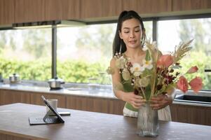 asiatico sorridente donna mettendo bellissimo fiori su vaso , godendo decorazione a casa foto