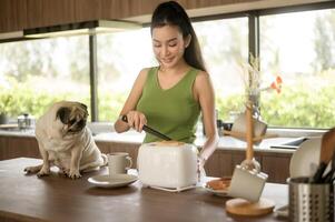 asiatico donna preparazione caffè e crostini pane per prima colazione godere con cane a il cucina tavolo nel il mattina foto
