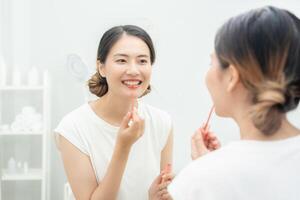 bellissimo asiatico donna sedersi nel davanti di un' specchio e Sorridi su trucco. viso di un' salutare donna l'applicazione trucco. annuncio, stile di vita , cosmetici, trucco Accessori, bellezza attività, estetista foto