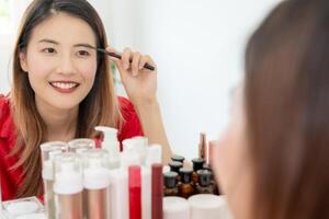 bellissimo asiatico donna sedersi nel davanti di un' specchio e Sorridi su trucco. viso di un' salutare donna l'applicazione trucco. annuncio, stile di vita , cosmetici, trucco Accessori, bellezza attività, estetista foto