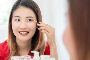 bellissimo asiatico donna sedersi nel davanti di un' specchio e Sorridi su trucco. viso di un' salutare donna l'applicazione trucco. annuncio, stile di vita , cosmetici, trucco Accessori, bellezza attività, estetista foto