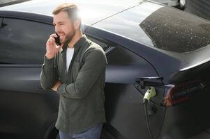 bello barbuto uomo nel casuale indossare, in piedi a il ricarica stazione e caricabatterie per un elettrico macchina. eco elettrico auto concetto foto