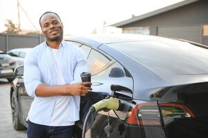 elegante africano uomo con coffe tazza nel mano inserti spina in il elettrico auto ricarica presa di corrente foto