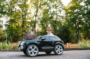 carino ragazzo nel equitazione un' nero elettrico auto nel il parco. divertente ragazzo cavalcate su un' giocattolo elettrico macchina. copia spazio. foto