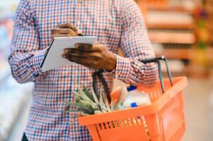 africano americano uomo in piedi nel supermercato la scelta prodotti mentre lettura Appunti a taccuino foto