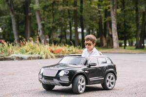 carino ragazzo nel equitazione un' nero elettrico auto nel il parco. divertente ragazzo cavalcate su un' giocattolo elettrico macchina. copia spazio. foto