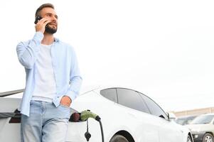 bellissimo giovane elegante uomo è con elettrico auto a giorno ricarica il veicolo foto