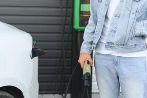 giovane bello uomo Tenere ricarica cavo a elettrico ricarica stazione punto in piedi vicino il suo nuovo auto foto