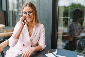 sorridente donna d'affari vestito nel formale indossare avendo piacevole conversazione su mobile Telefono con amico, contento attraente femmina A proposito di su cellula telefono mentre riposo nel ufficio interno dopo opera foto