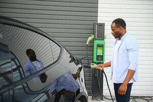 africano uomo in piedi vicino elettrico auto con ricarica cavo nel tappo. eco amichevole veicolo ricarica su stazione. stili di vita concetto foto