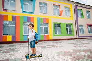 adolescenziale ragazzo con calcio scooter vicino moderno scuola. bambino con zaino e libro all'aperto. inizio di Lezioni. primo giorno di autunno. indietro per scuola. foto