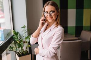 attività commerciale donna su il Telefono a ufficio. giovane attività commerciale donna avendo Telefono chiamata a il ufficio. sorridente attività commerciale donna. bella giovane attività commerciale utilizzando smartphone a il soffitta ufficio. foto