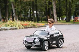 carino ragazzo nel equitazione un' nero elettrico auto nel il parco. divertente ragazzo cavalcate su un' giocattolo elettrico macchina. copia spazio. foto