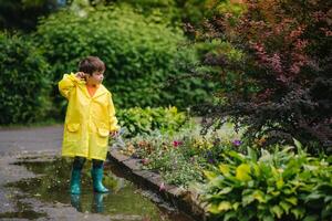 poco ragazzo giocando nel piovoso estate parco. bambino con ombrello, impermeabile cappotto e stivali salto nel pozzanghera e fango nel il pioggia. ragazzo a piedi nel estate pioggia all'aperto divertimento di qualunque tempo atmosferico. contento infanzia. foto