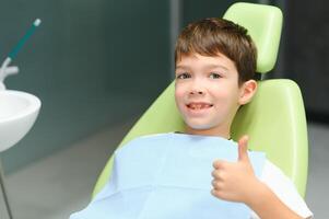 poco ragazzo si siede su dal dentista sedia nel bene umore dopo dentale procedure. giovane paziente con salutare denti foto