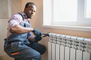 uomo nel abbigliamento da lavoro tuta da lavoro utilizzando utensili mentre installazione o riparazione riscaldamento termosifone nel camera foto