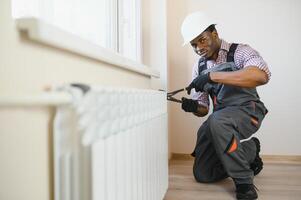 afro riparatore nel tuta da lavoro utilizzando utensili mentre installazione o riparazione riscaldamento termosifone foto