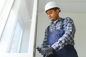 indiano operaio nel tuta da lavoro installazione o regolazione plastica finestre nel il vivente camera a casa foto