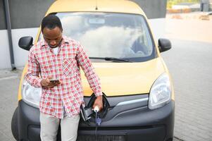 africano americano uomo sta Il prossimo per elettrico consegna furgoni a elettrico veicolo ricarica stazione foto