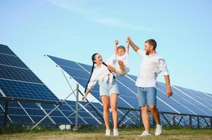 un' largo tiro di un' contento famiglia in piedi insieme e sorridente a telecamera con un' grande solare pannello nel sfondo foto