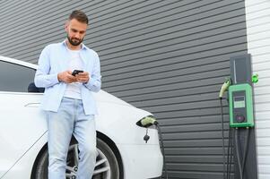 bellissimo giovane elegante uomo è con elettrico auto a giorno ricarica il veicolo foto