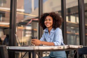 bellissimo giovane africano donna Tenere caffè tazza seduta al di fuori bar con il computer portatile Iscritto con un altro foto