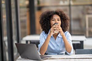 giovane afroamericano donna Tenere caffè tazza seduta al di fuori bar con il computer portatile. sollevato un' tazza di caffè e Bevuto e fissava lontano. foto