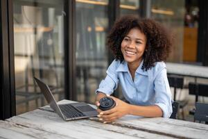 giovane afroamericano donna Tenere caffè tazza seduta al di fuori bar con il computer portatile. Guarda su per interessante cose e Sorridi a il tuo dintorni. foto