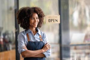 riuscito africano americano donna nel grembiule in piedi caffè negozio porta. contento piccolo attività commerciale proprietario. sorridente ritratto di imprenditore in piedi con copia spazio. foto