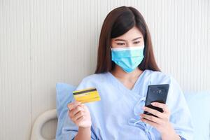 asiatico femmina paziente indossare maschera nel ospedale paziente camera lei detiene un' smartphone e un' credito carta per pagare in linea. concetto di medico Servizi. pazienti infetto con coronavirus. Salute assicurazione foto