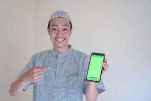 musulmano asiatico uomo felice, sorridente quando mostrare il smartphone con verde schermo quando Ramadan celebrazione. il foto è adatto per uso per Ramadhan manifesto e musulmano soddisfare media.