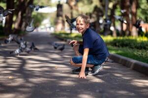 un' ragazzo nel un' blu maglietta si siede su il asfalto nel il parco e sembra a il piccioni. foto