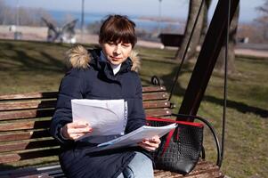 un' giovane femmina contabile si siede su un' parco panchina e sembra attraverso segnalazione documenti durante sua rompere foto