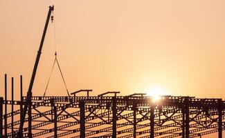 silhouette gru sollevamento metallo castellato fascio per installazione su grande industriale edificio struttura nel costruzione luogo la zona contro tramonto cielo sfondo foto